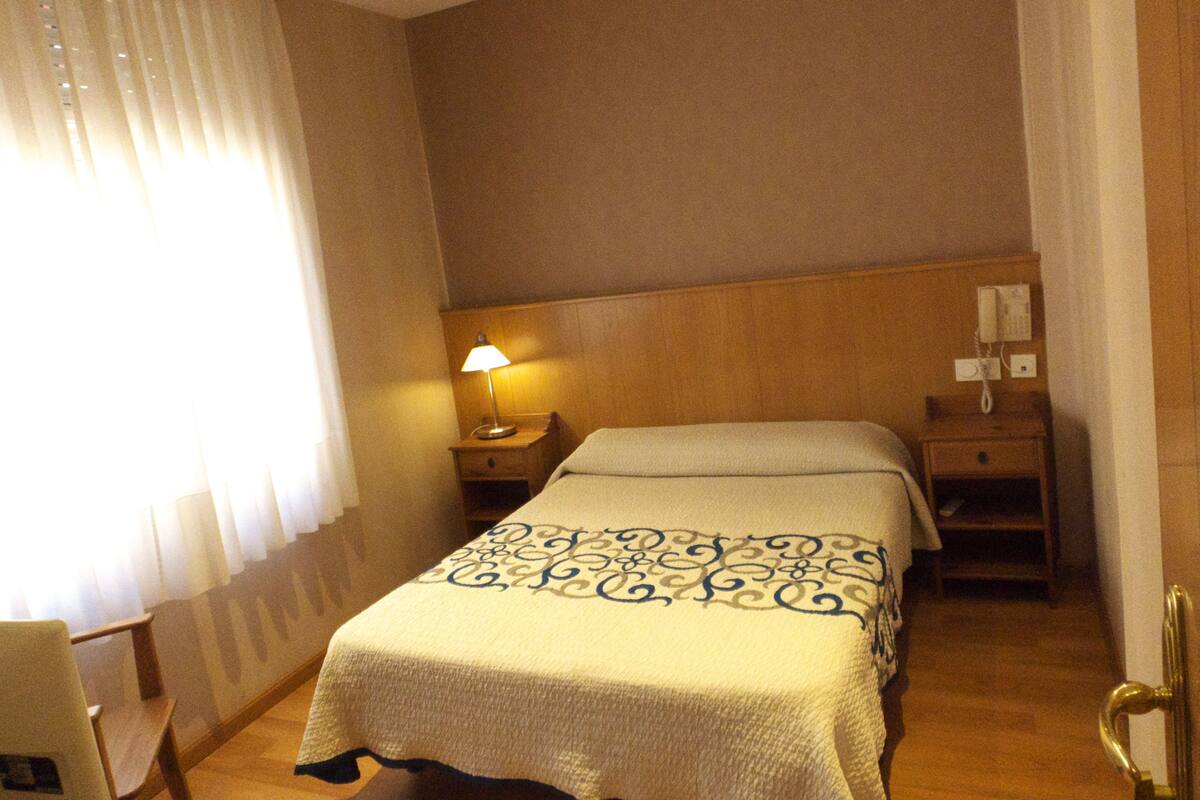 Quarto de hotel com cama, mesa de cabeceira com abajur, cabeceira de madeira, telefone e janela com cortinas.