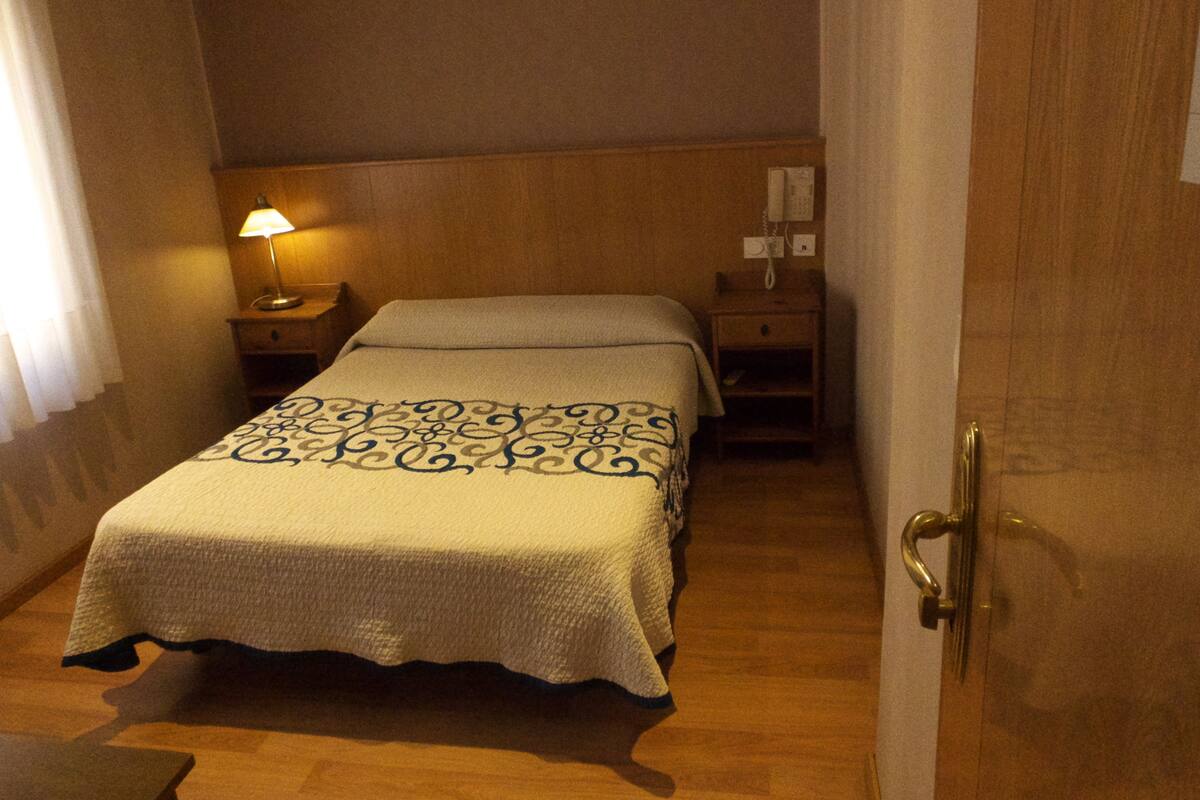 Quarto de hotel com uma cama, mesa de cabeceira, luminária e porta de madeira.
