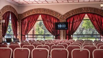 A conference room with rows of red chairs, brick walls, and large windows with red curtains. A digital clock displays 10:00.