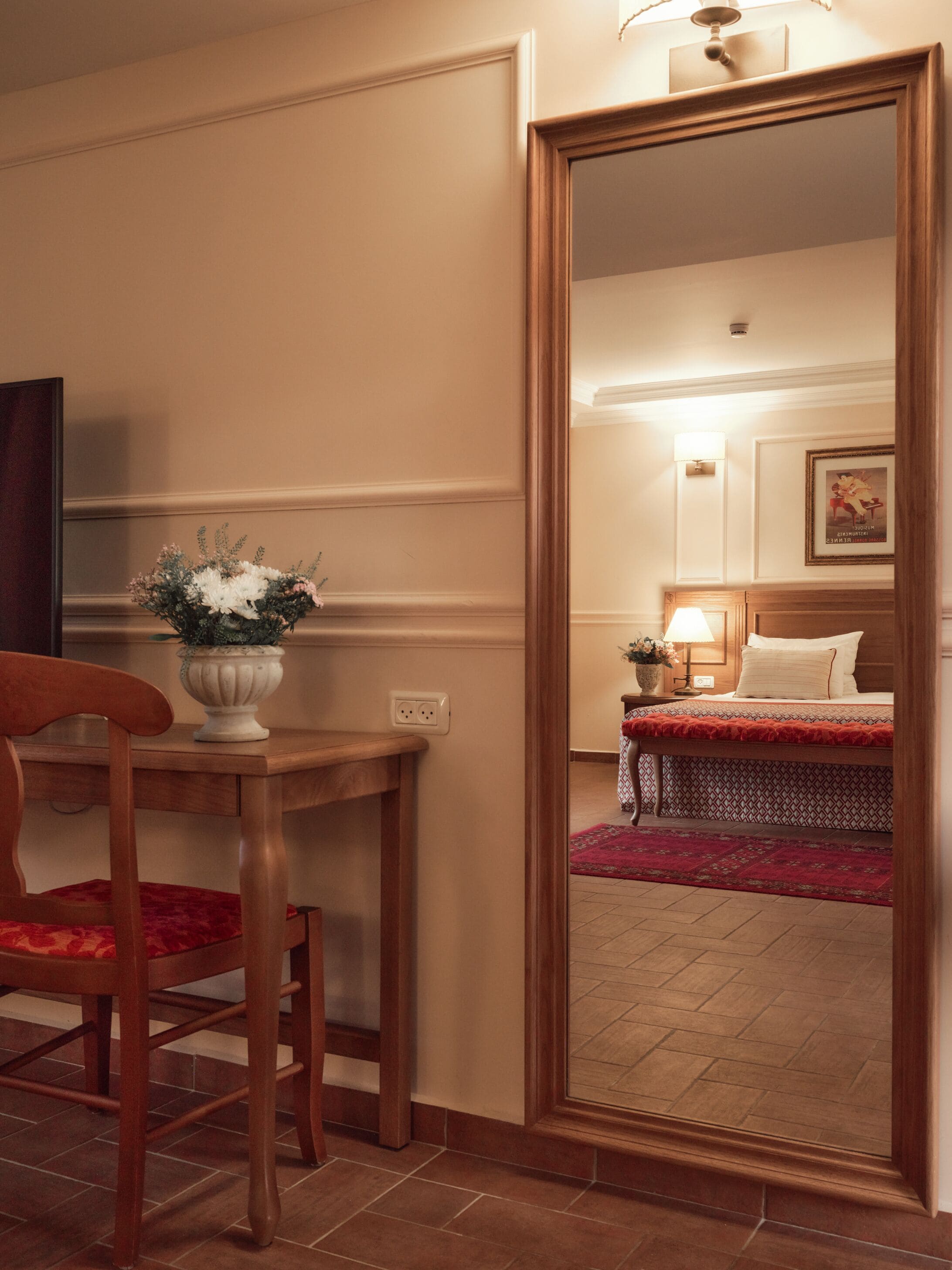 Ein Hotelzimmer mit Holzstuhl, Tisch mit Blumenvase, Bett mit roter Bettdecke und Spiegel, der den Raum reflektiert.