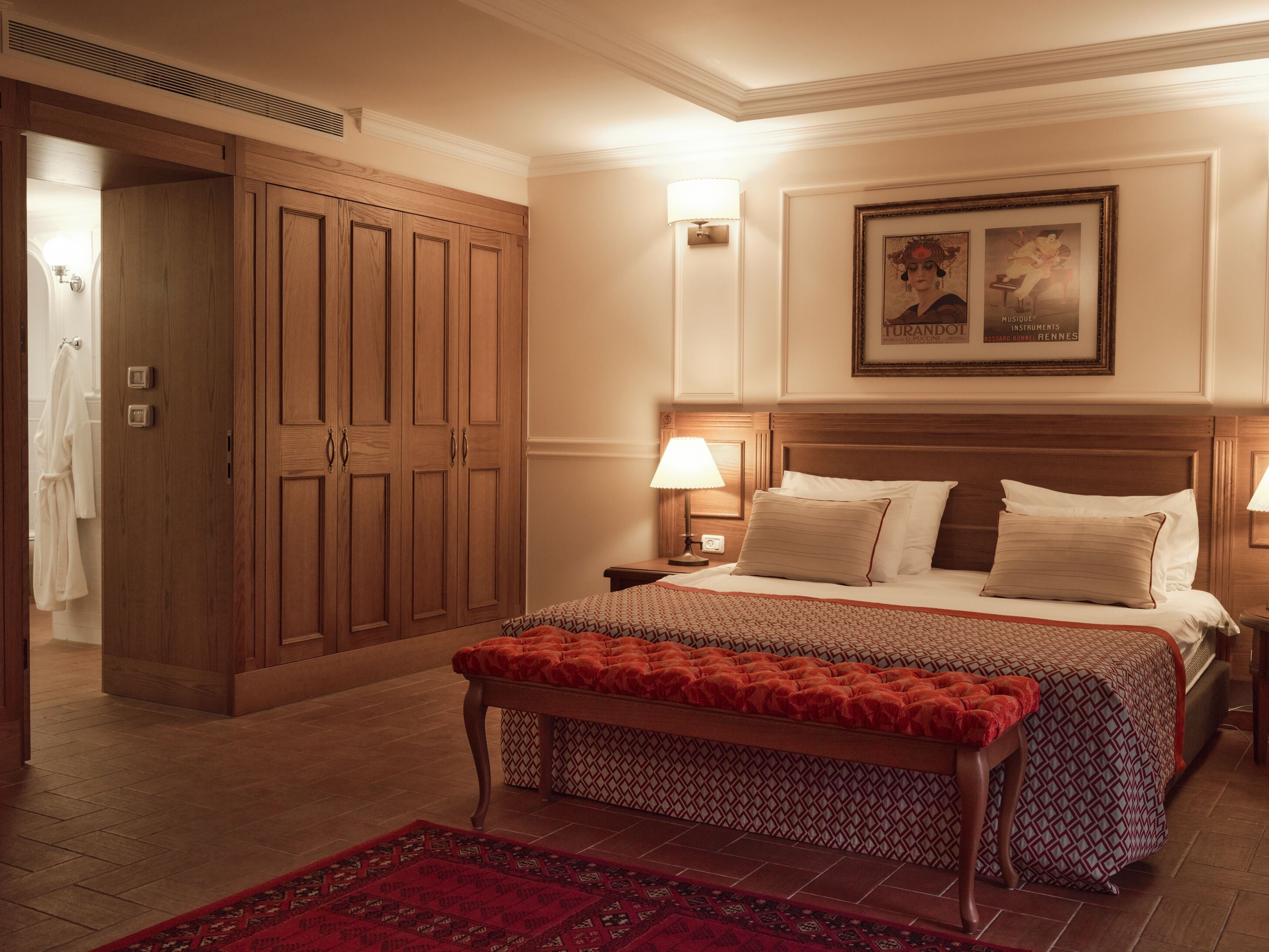 Una habitación de hotel con un armario de madera grande, una cama con dos almohadas, una mesita de noche con una lámpara y una alfombra roja estampada.