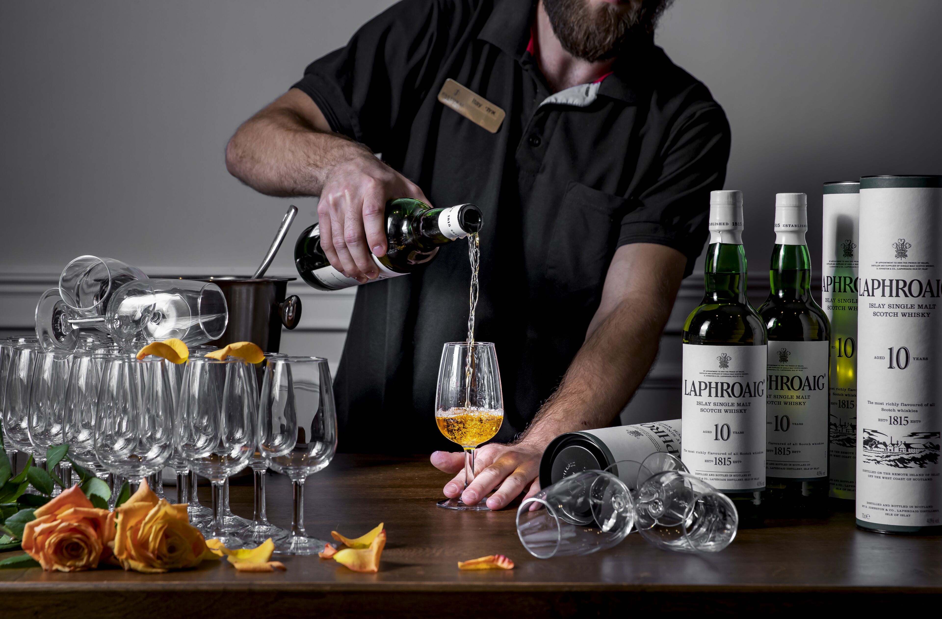 A person pouring a drink into a glass, surrounded by empty glasses and bottles of Laphroaig whisky.