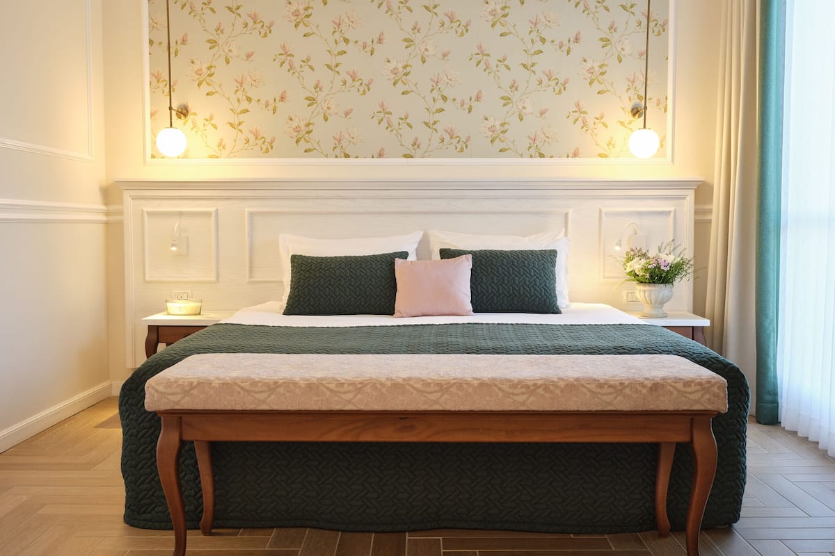 Un lit bien fait, recouvert d’une couverture verte et agrémenté d’oreillers, un banc en bois au pied du lit, une table de chevet avec une lampe, et un mur orné d’un papier peint à motifs floraux.
