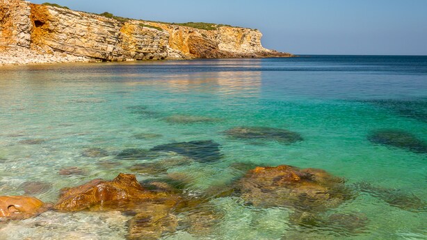 A coastal scene with clear turquoise water, rocky formations, and a cliff with vegetation.