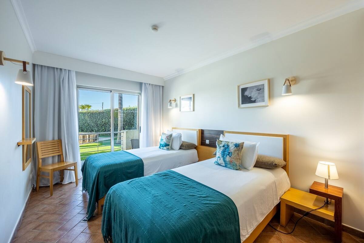 A hotel room with two beds, a wooden chair, a small table with a lamp, and a balcony with a view of greenery.