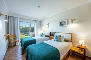 A hotel room with two beds, a wooden chair, a small table with a lamp, and a balcony with a view of greenery. - Pontalaia Apartamentos Turisticos (Vila do Bispo)