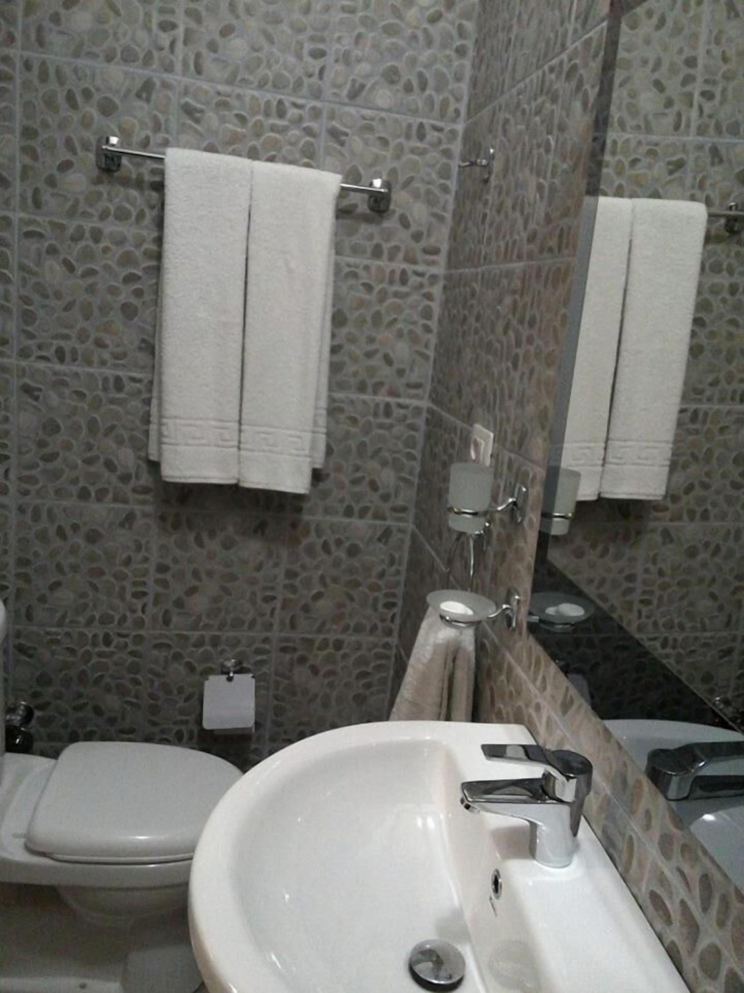 Un baño con un lavabo blanco, un inodoro y un espejo. Hay dos toallas blancas colgadas en toalleros. Las paredes presentan un diseño que imita a la piedra.