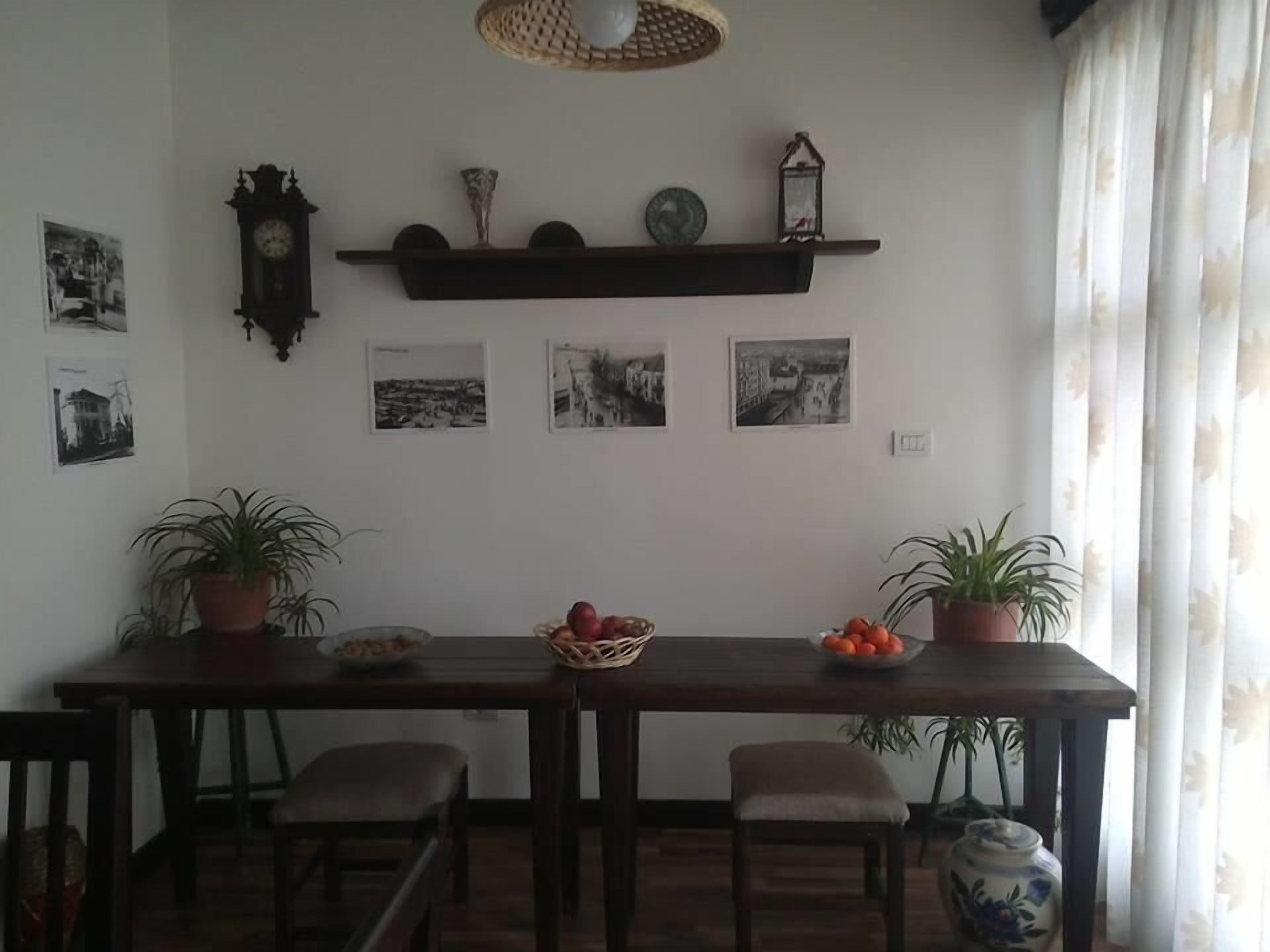 Un comedor con una mesa de madera, cuatro sillas, dos macetas con plantas, un frutero y un estante de pared con adornos.