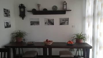 Un comedor con una mesa de madera, cuatro sillas, dos macetas con plantas, un frutero y un estante de pared con adornos.