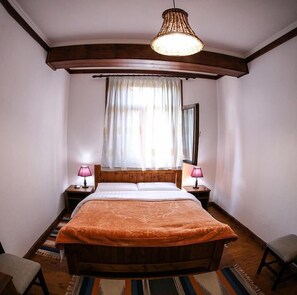 Une chambre à coucher avec un lit en bois, deux lampes de chevet, une grande fenêtre avec des rideaux légers et un luminaire suspendu.