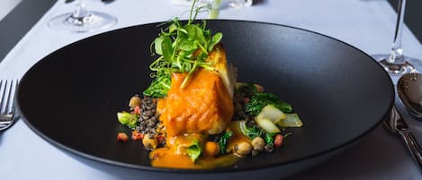 Un plato negro con un plato de pescado, vegetales verdes y verduras sobre una mesa con mantel blanco.