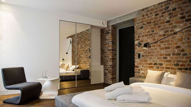 Una habitación de hotel moderna con pared de ladrillo, una cama grande con ropa blanca, un sillón gris, una mesita blanca pequeña con vasos y un espejo que refleja la habitación.