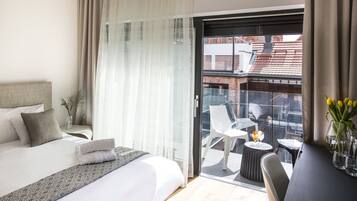 A hotel room with a bed, a small table with a drink, a chair, and a view of the city through a glass door.