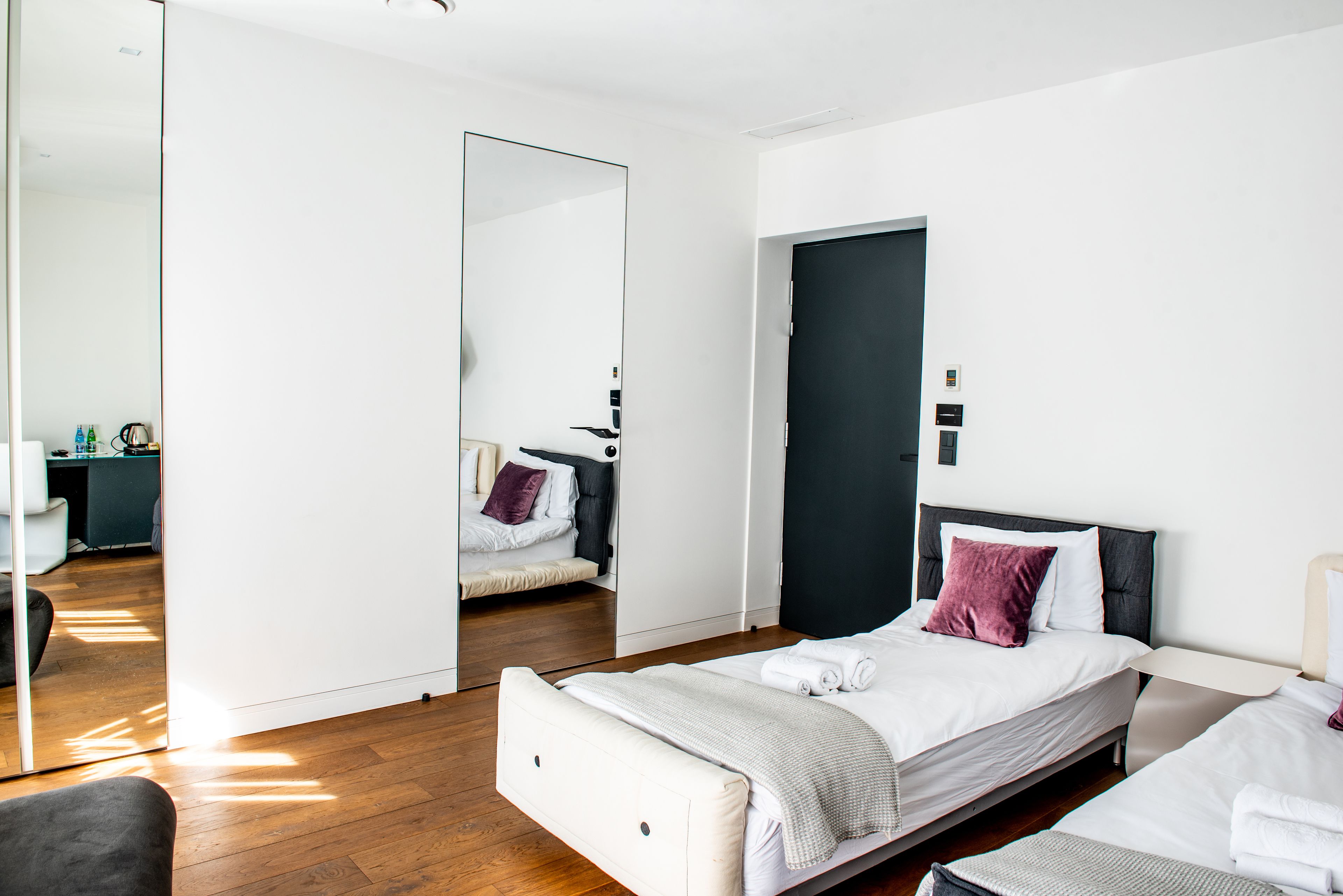 Una habitación de hotel moderna con un espejo grande, una cama con ropa de cama blanca, una puerta oscura y un escritorio pequeño con una silla.