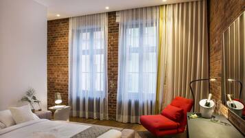 Quarto de hotel moderno com parede de tijolos, grandes janelas com cortinas leves, cama com roupa de cama branca, poltrona vermelha e mesa de cabeceira com abajur.