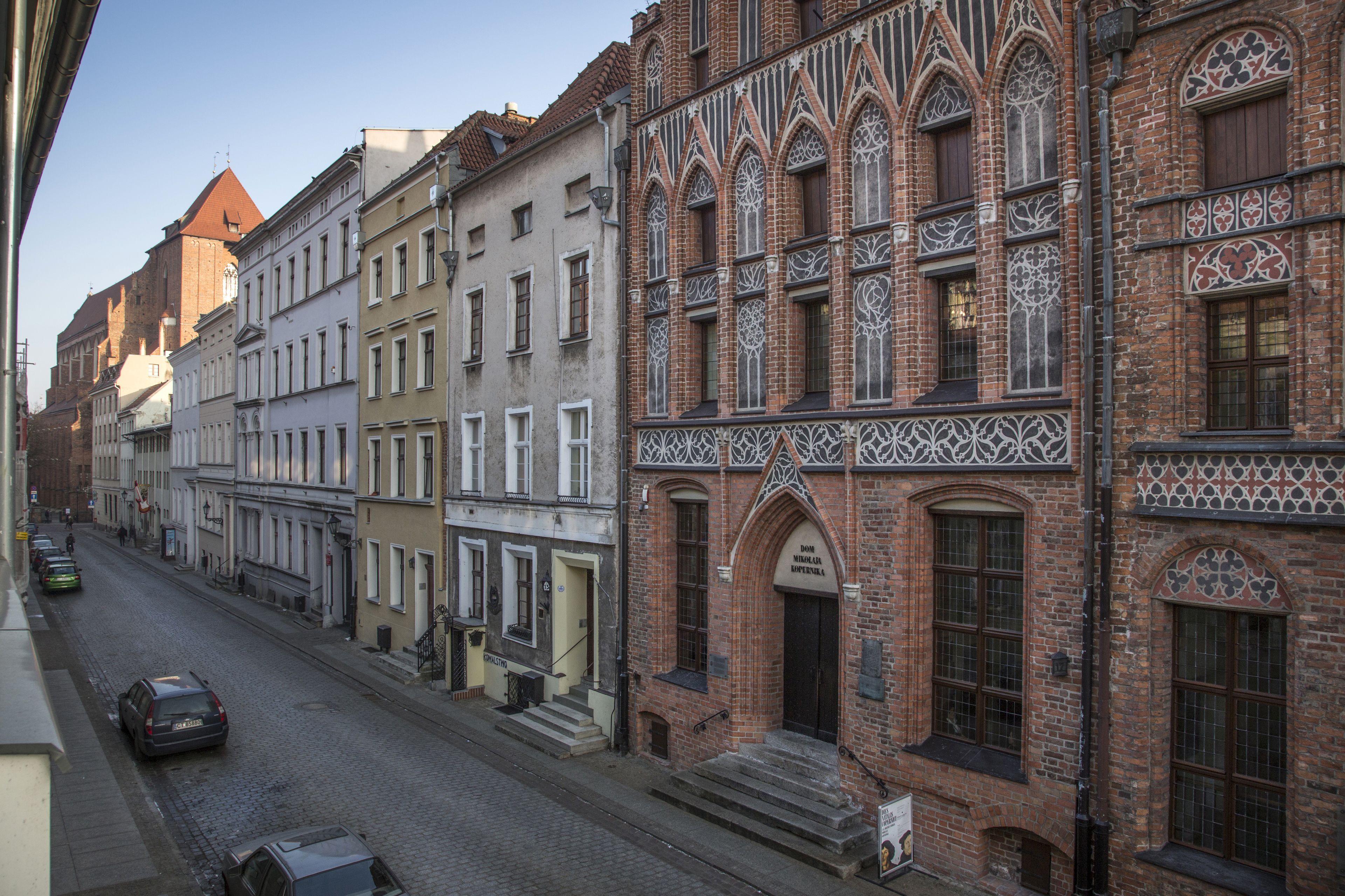 A street with historic buildings, including a building with a pointed arch and intricate brickwork.