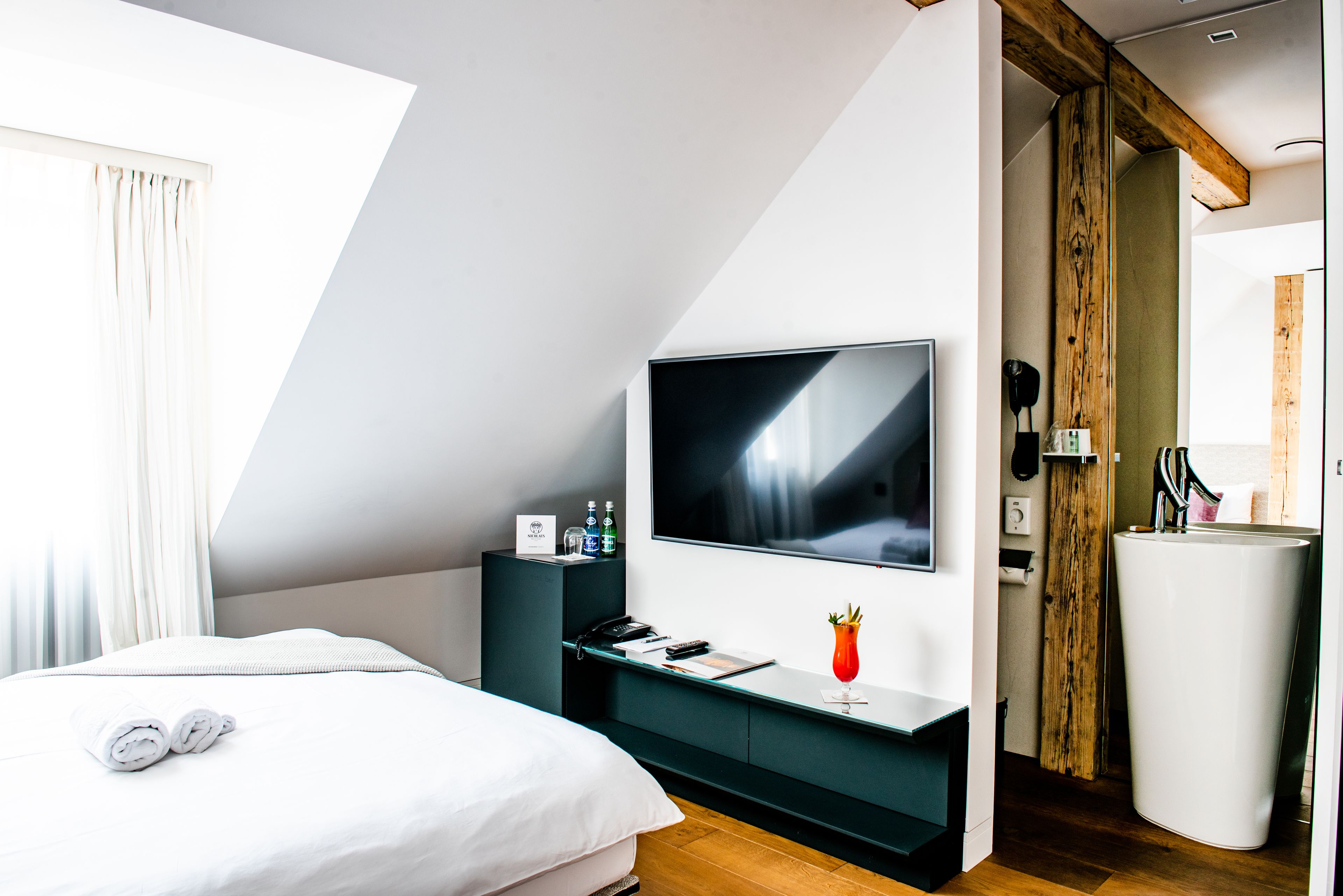 A modern hotel room with a large bed, a flat-screen TV, a small desk with a phone and a glass of drink, and a wooden accent wall.