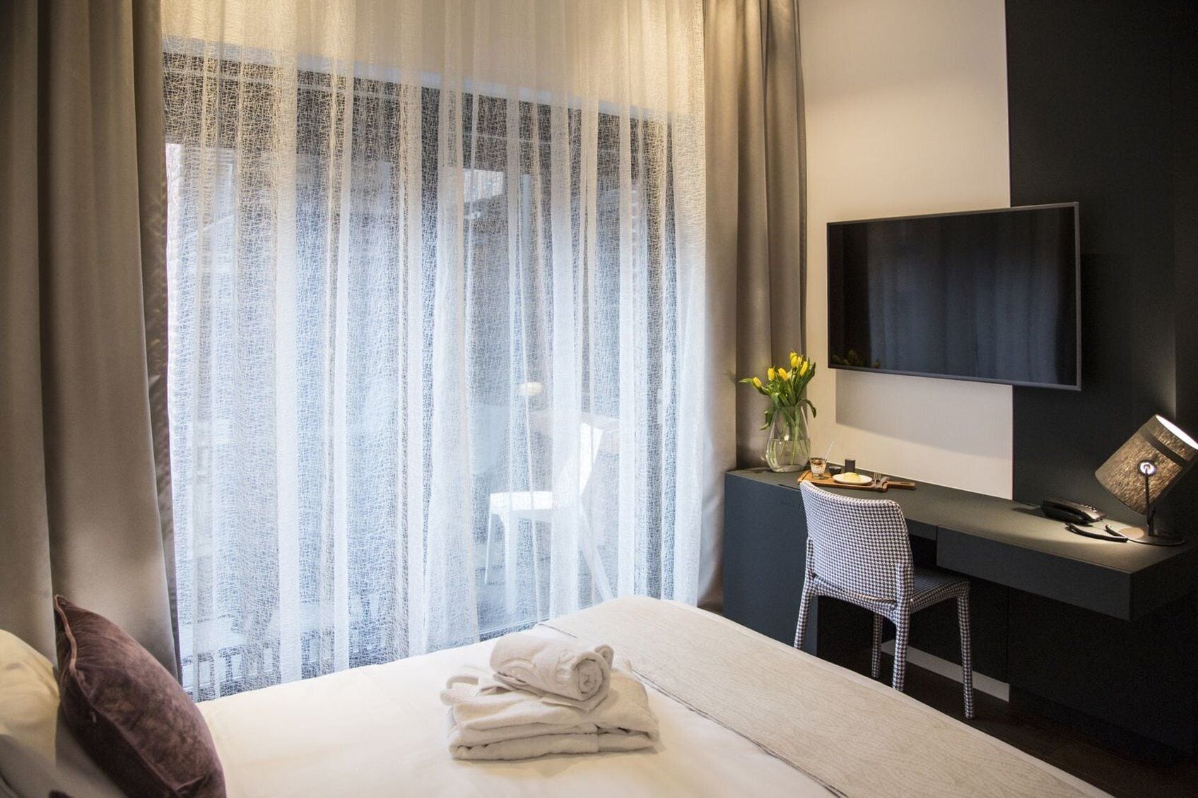 A hotel room with a bed, a desk, a chair, a TV, and a window with sheer curtains.