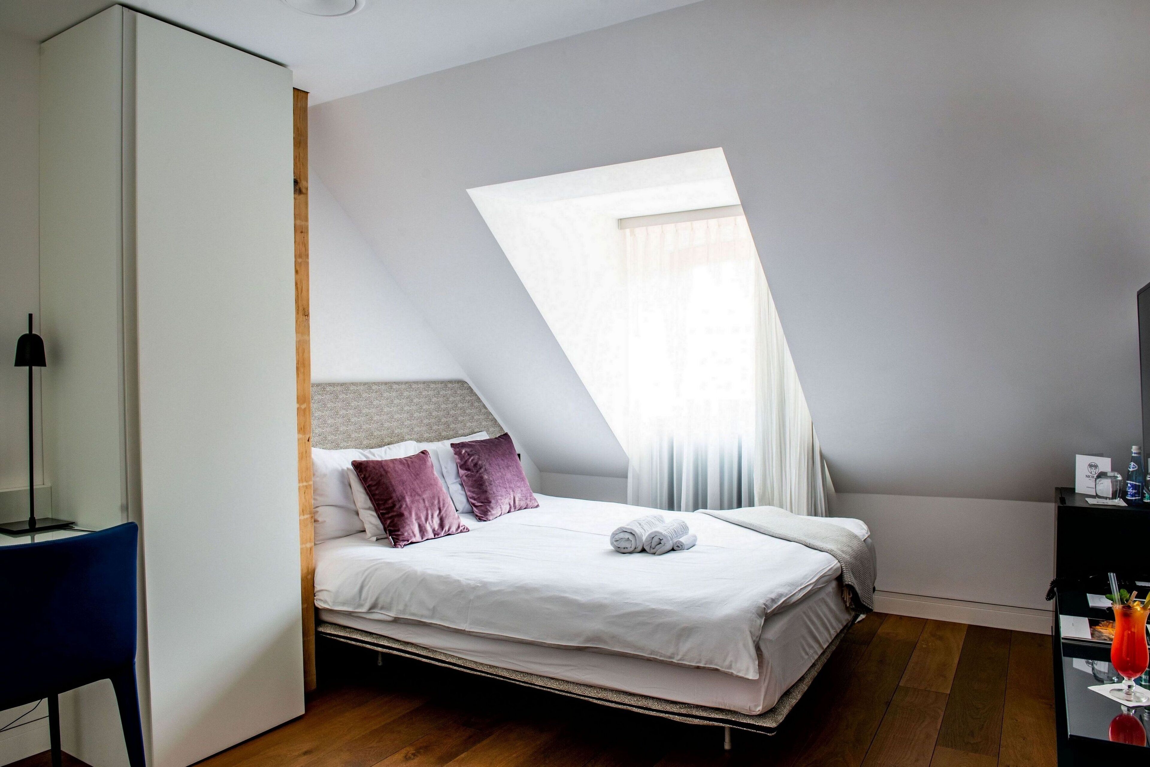A modern bedroom with a sloped ceiling, a bed with white bedding and purple pillows, a desk with a chair, and a window with sheer curtains.