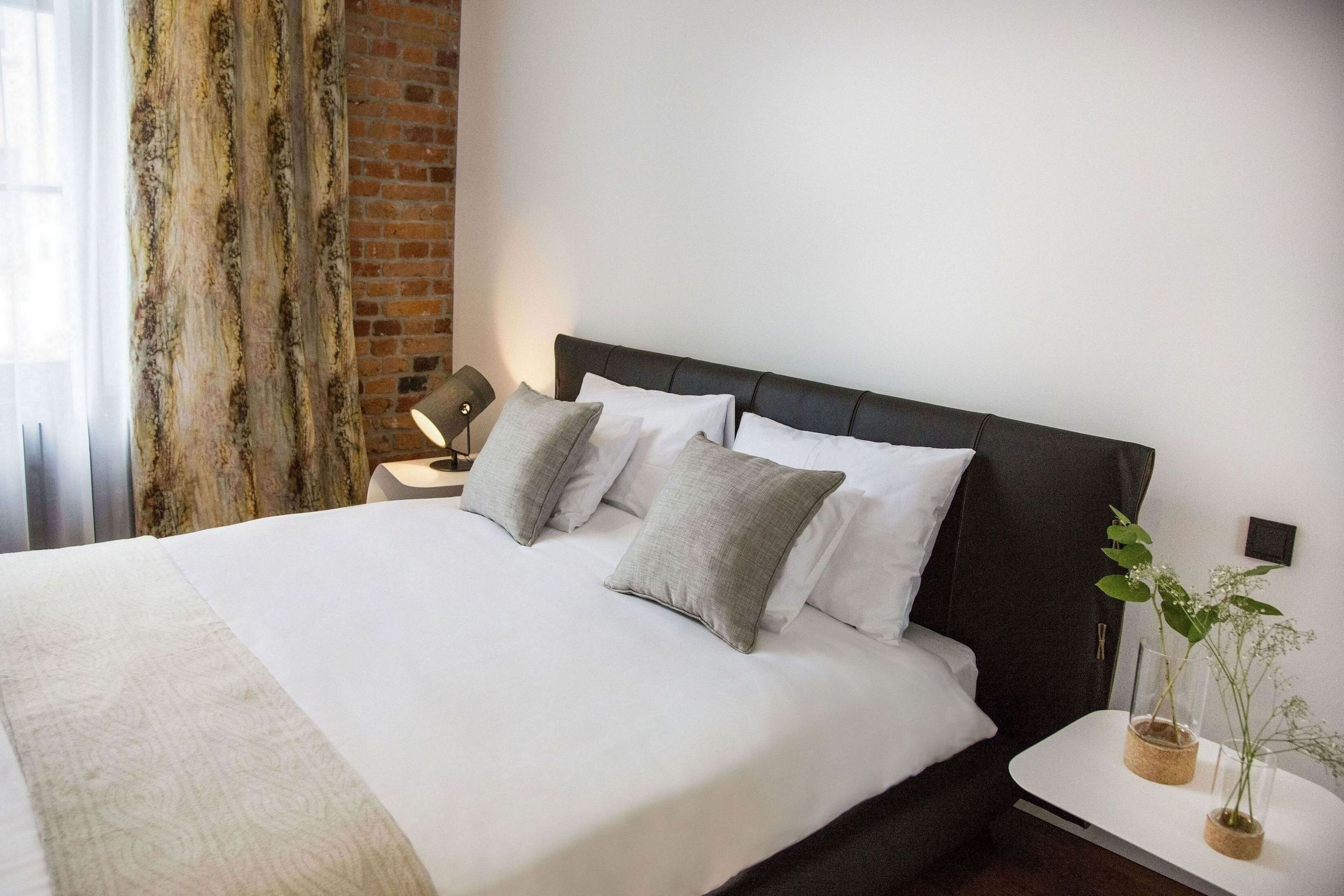 Una cama bien tendida con sábanas blancas y almohadas grises, una mesita de noche con lámpara, y una planta en maceta sobre una pequeña mesa junto a la cama.