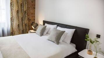 En pænt redt seng med hvide sengetæpper og grå puder, et natbord med en lampe, og en potteplante på et lille bord ved siden af sengen.
