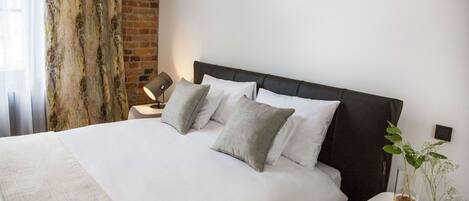 Una cama bien tendida con sábanas blancas y almohadas grises, una mesita de noche con lámpara, y una planta en maceta sobre una pequeña mesa junto a la cama.