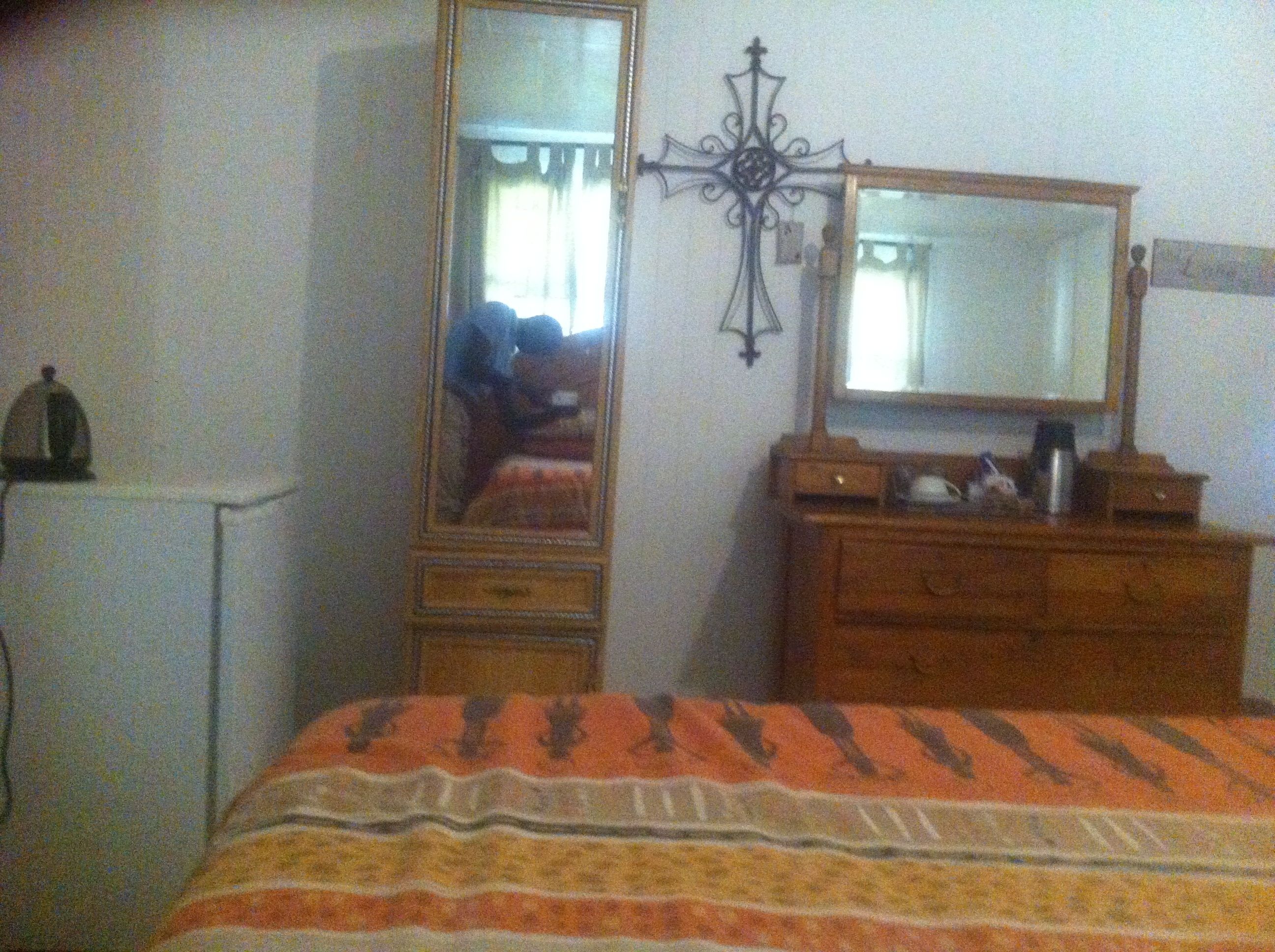 Une chambre à coucher comprenant un lit, une commode, un miroir et une petite table avec une bouilloire.