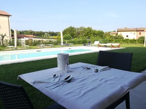 Une table à manger installée à l’extérieur, avec vue sur la piscine, entourée de verdure et de bâtiments.