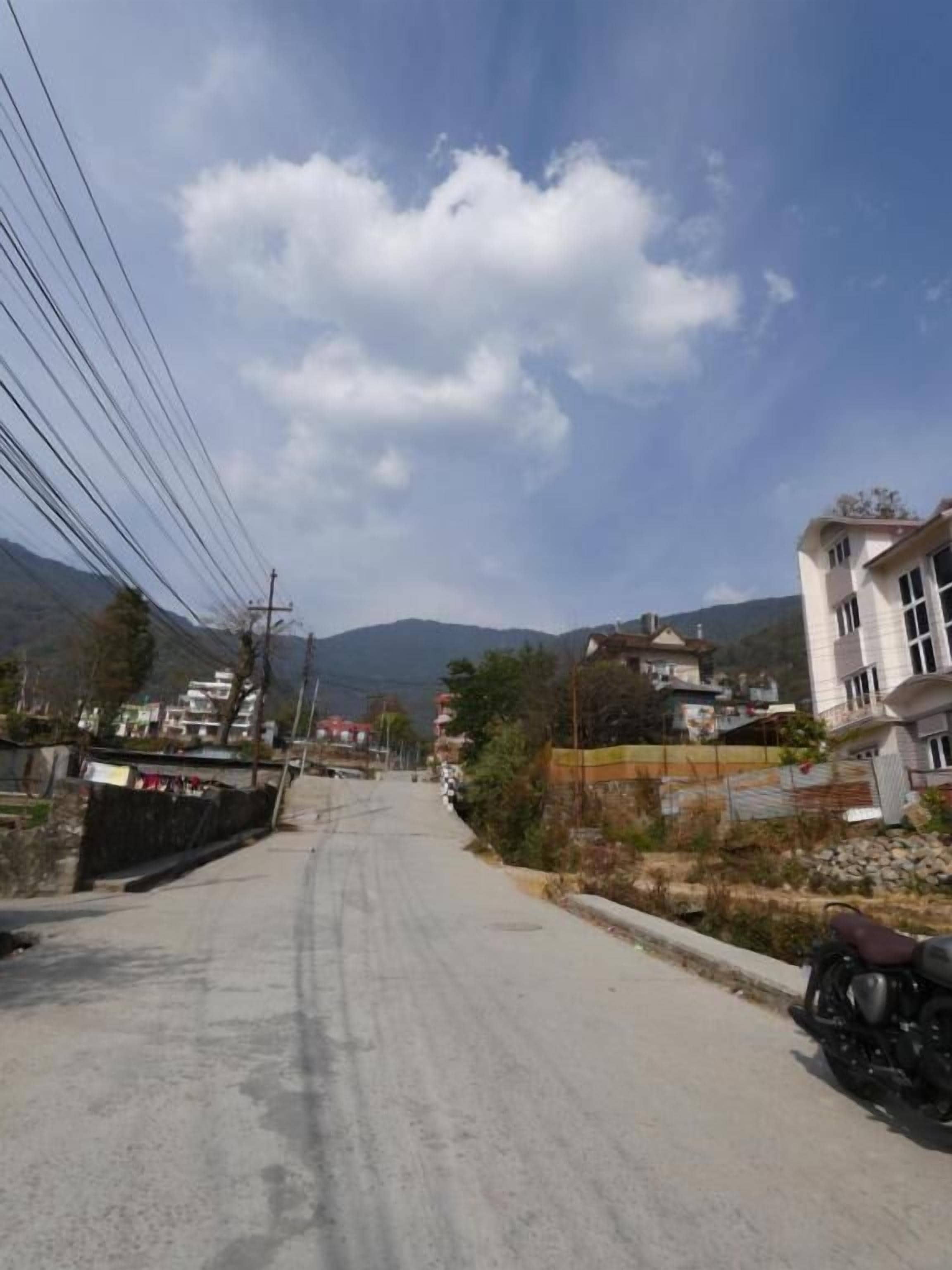 A paved road with a motorcycle parked on the right, surrounded by residential buildings and mountains in the background.