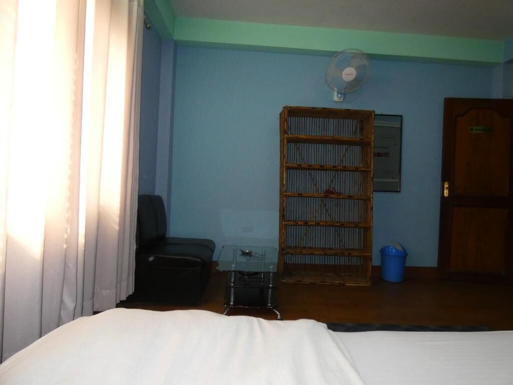 A hotel room with a bed, a small table, a fan, a bookshelf, and a door.
