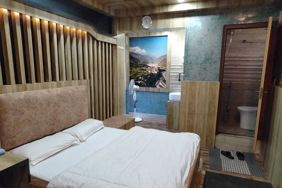 Un dormitorio moderno con una cama grande, cabecera de madera, mesita de noche pequeña y un baño visible a través de una puerta abierta.