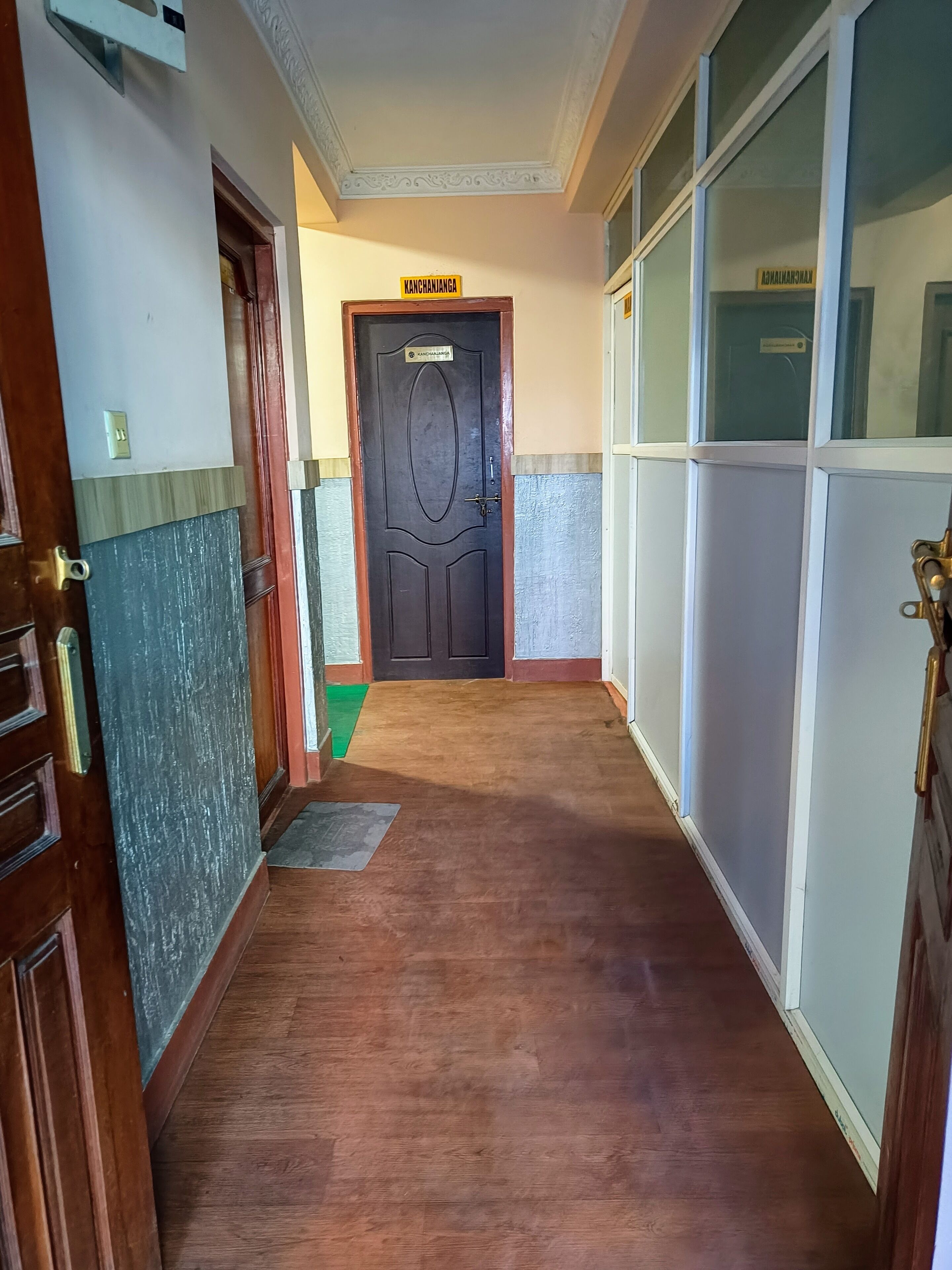 A hallway with a wooden door labeled "KANCHANJANGA" and a glass door with a sign.