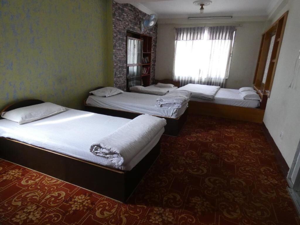 A hotel room with four single beds, a bookshelf, a window with curtains, and a patterned carpet.