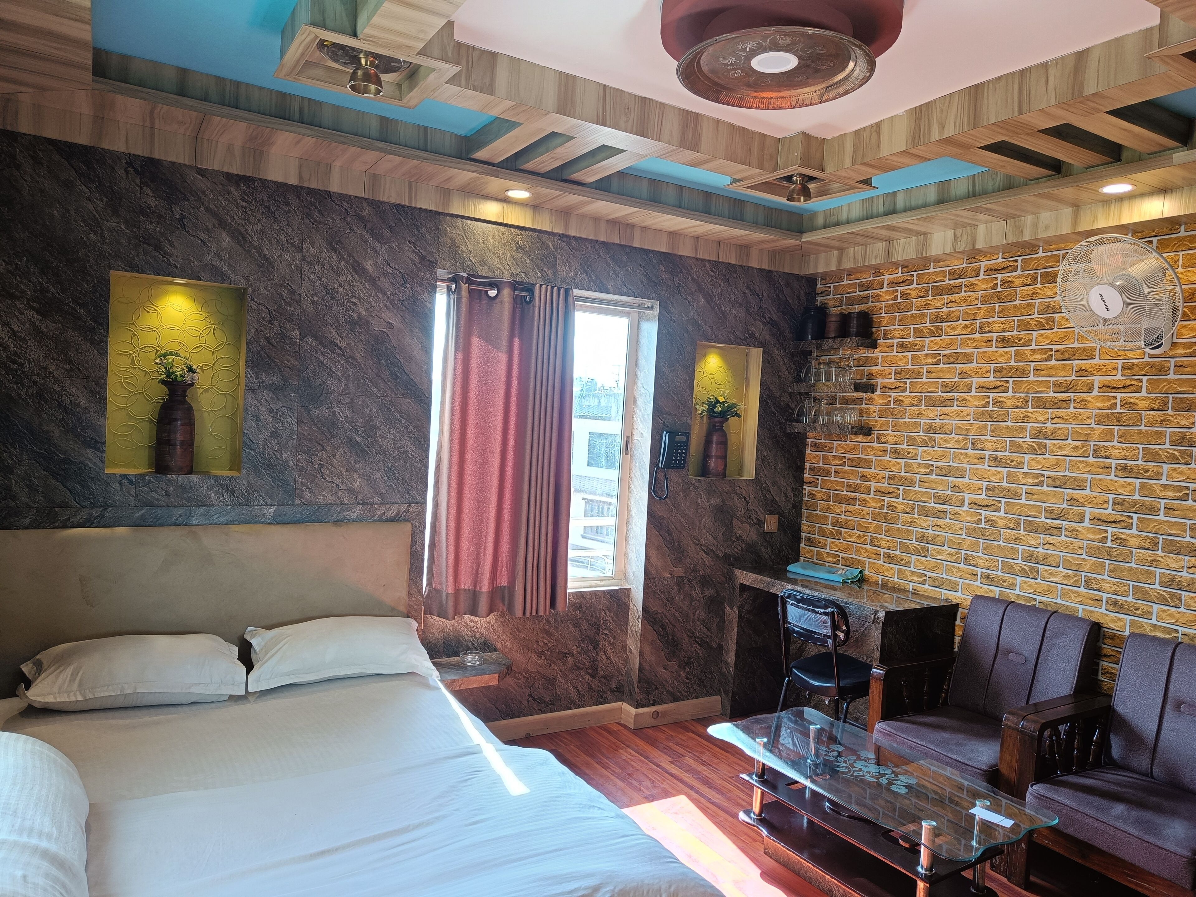 A modern hotel room with a large bed, a glass coffee table, a brick-patterned wall, and a ceiling fan.