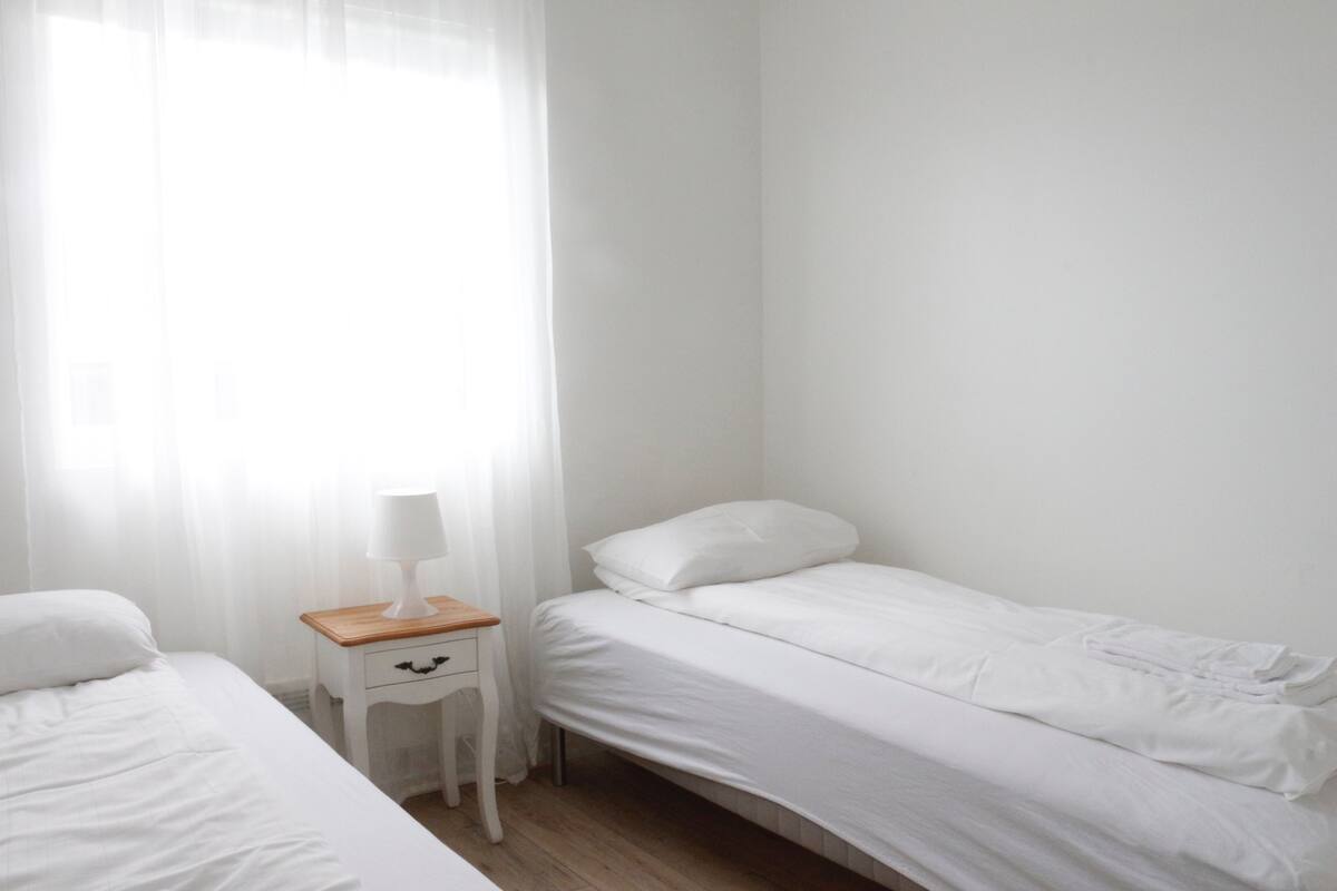 A small, clean room with a single bed, a nightstand with a lamp, and a window with sheer curtains.