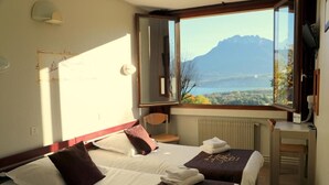 Habitación de hotel con dos camas, un escritorio y un amplio ventanal con vistas a la montaña.
