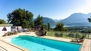Una piscina con sillas de descanso y sombrillas, rodeada por una terraza de madera, con vistas a la montaña al fondo.