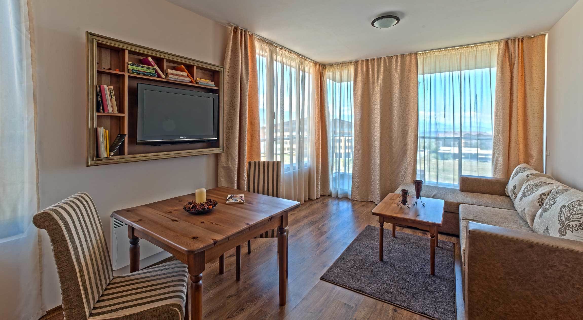 A living room with a wooden table, striped chairs, a sofa, a TV mounted on the wall, and large windows with curtains.