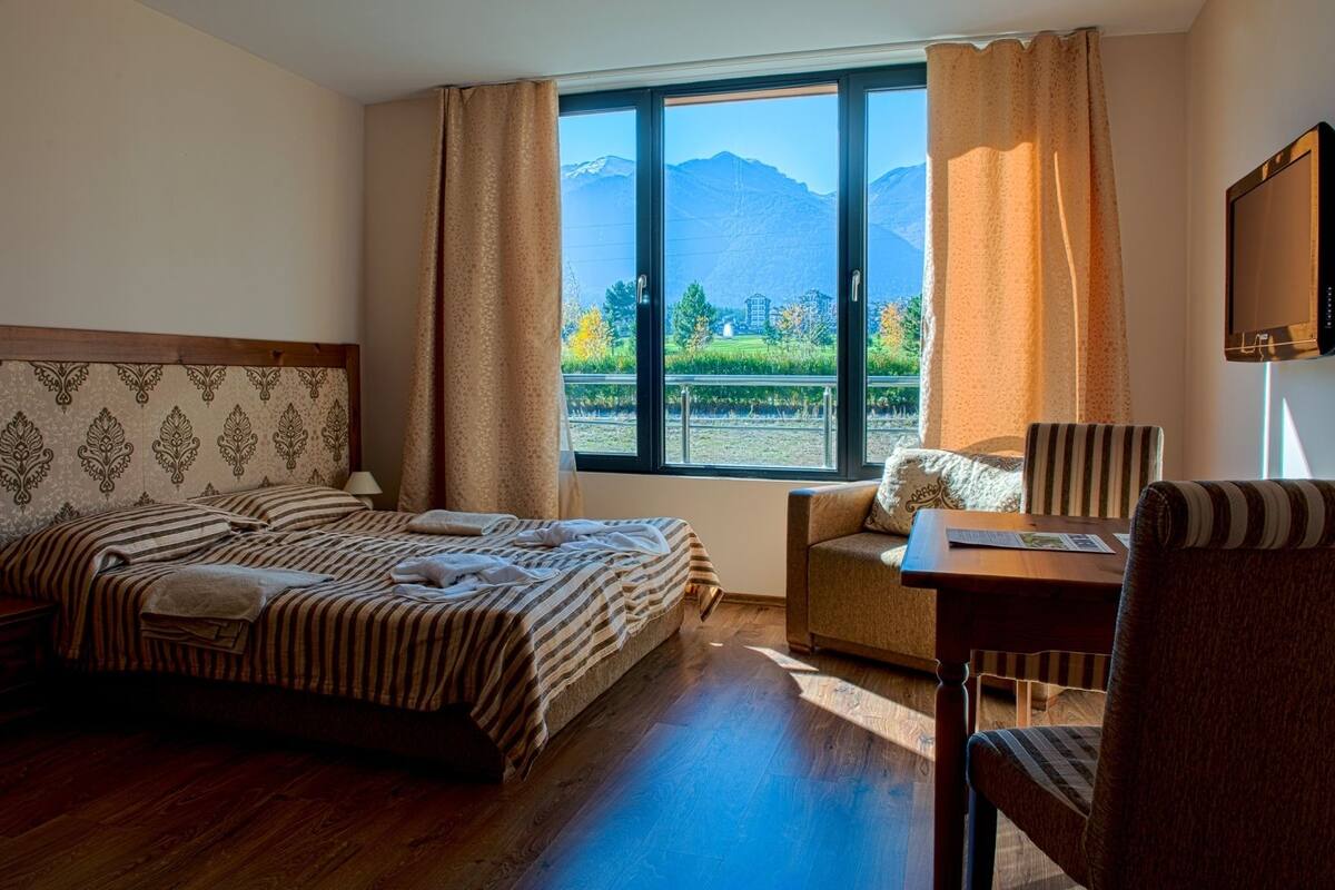 A hotel room with a bed, a chair, a desk, and a large window with a mountain view.