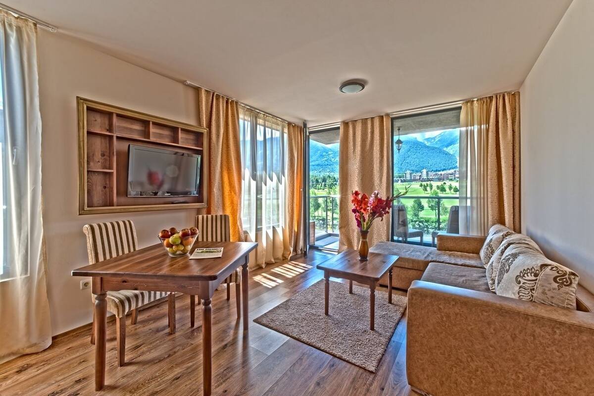 A spacious living room with a wooden dining table, a sofa, and a TV mounted on a wooden panel. Large windows offer a view of a mountainous landscape.