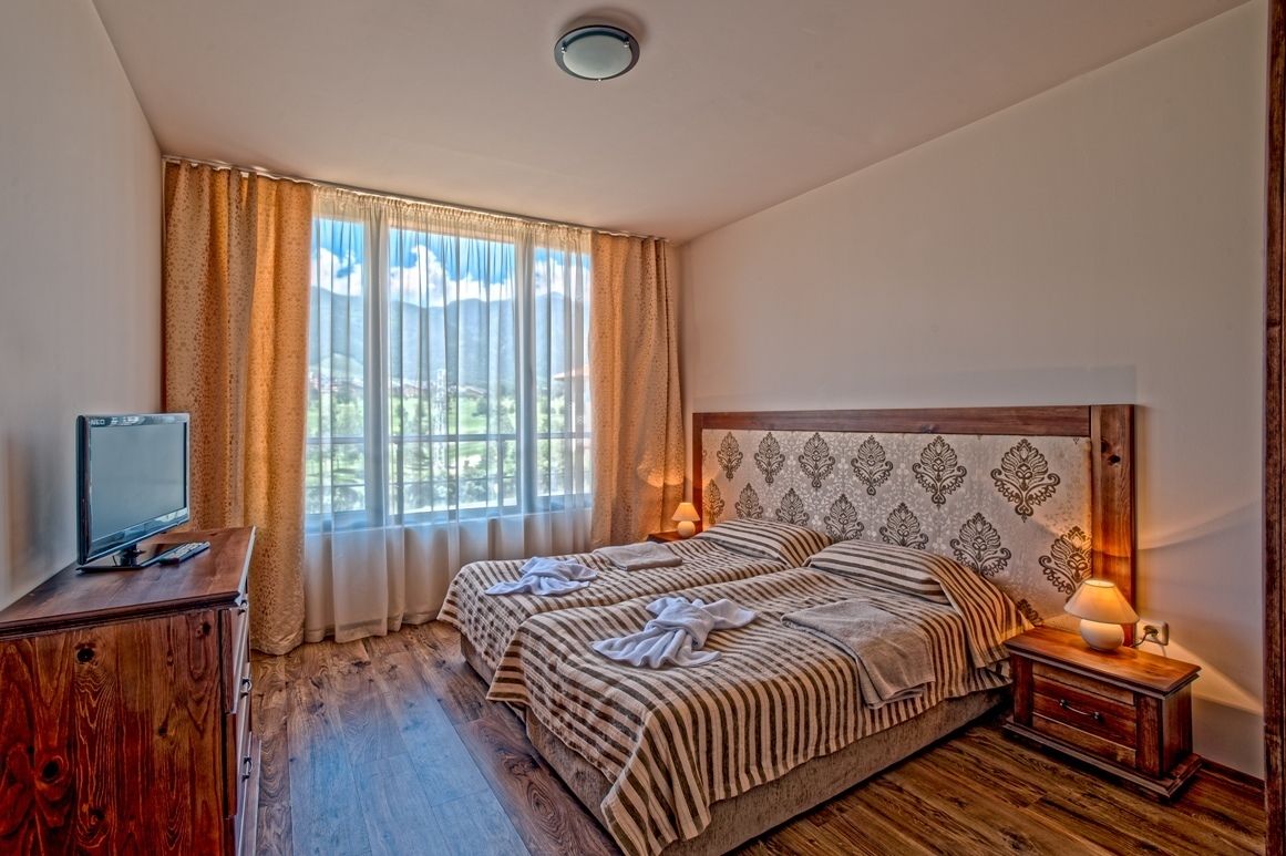 A bedroom with two beds, a TV on a wooden cabinet, a nightstand with a lamp, and a window with curtains.