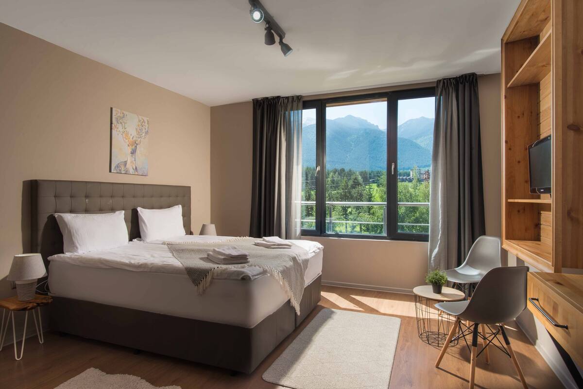A modern hotel room with a large bed, a desk with two chairs, a television, and a window with a mountain view.