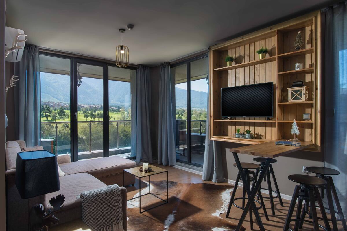 A modern living room with a large TV, a sofa, a coffee table, and a dining area with stools.
