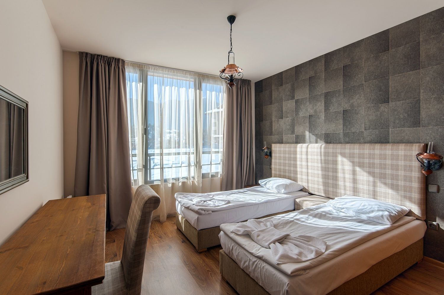 Ein Hotelzimmer mit zwei Betten, einem Holztisch, einem Stuhl, einem großen Fenster mit Vorhängen und einer Wandlampe.