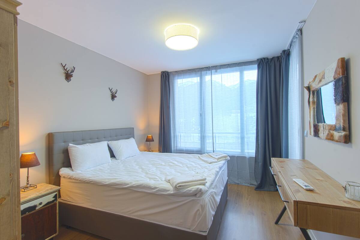 A hotel room with a large bed, bedside tables, a desk, and a window with curtains.