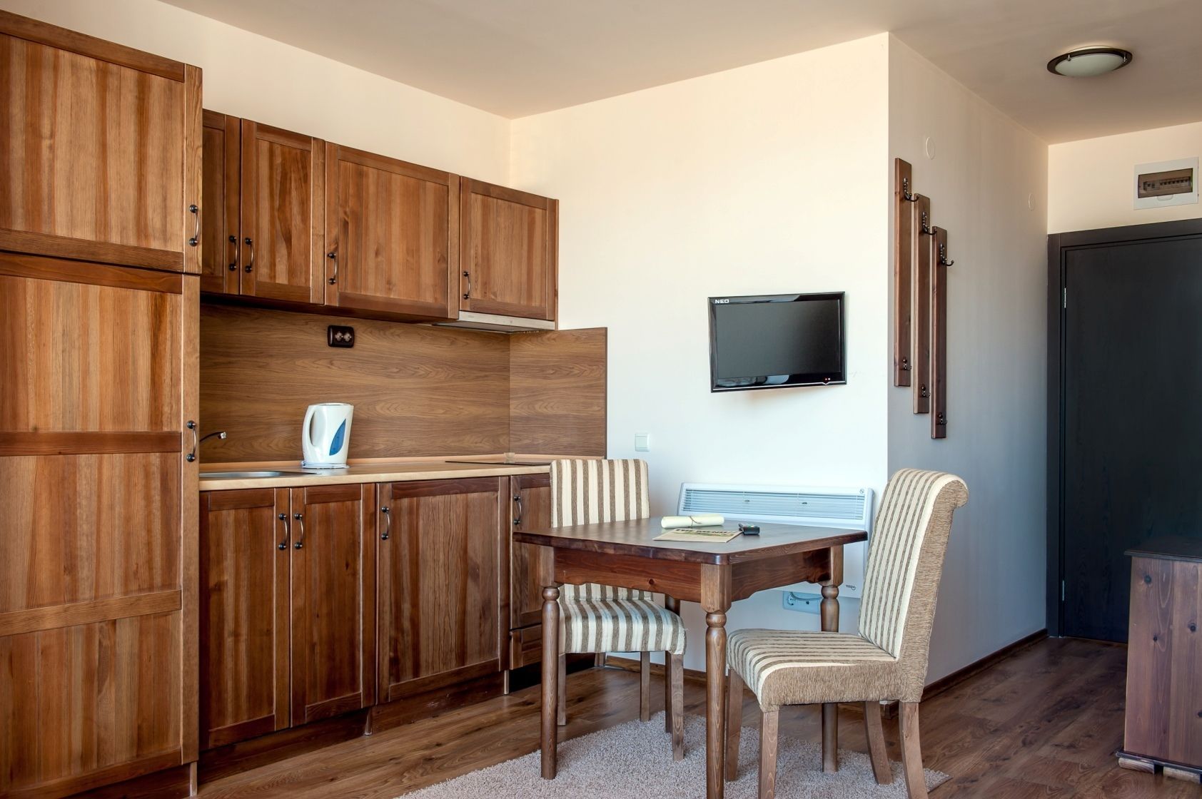 Una cocina compacta con armarios de madera, un microondas integrado, una mesa de comedor con dos sillas a rayas y un televisor de pantalla plana montado en la pared.