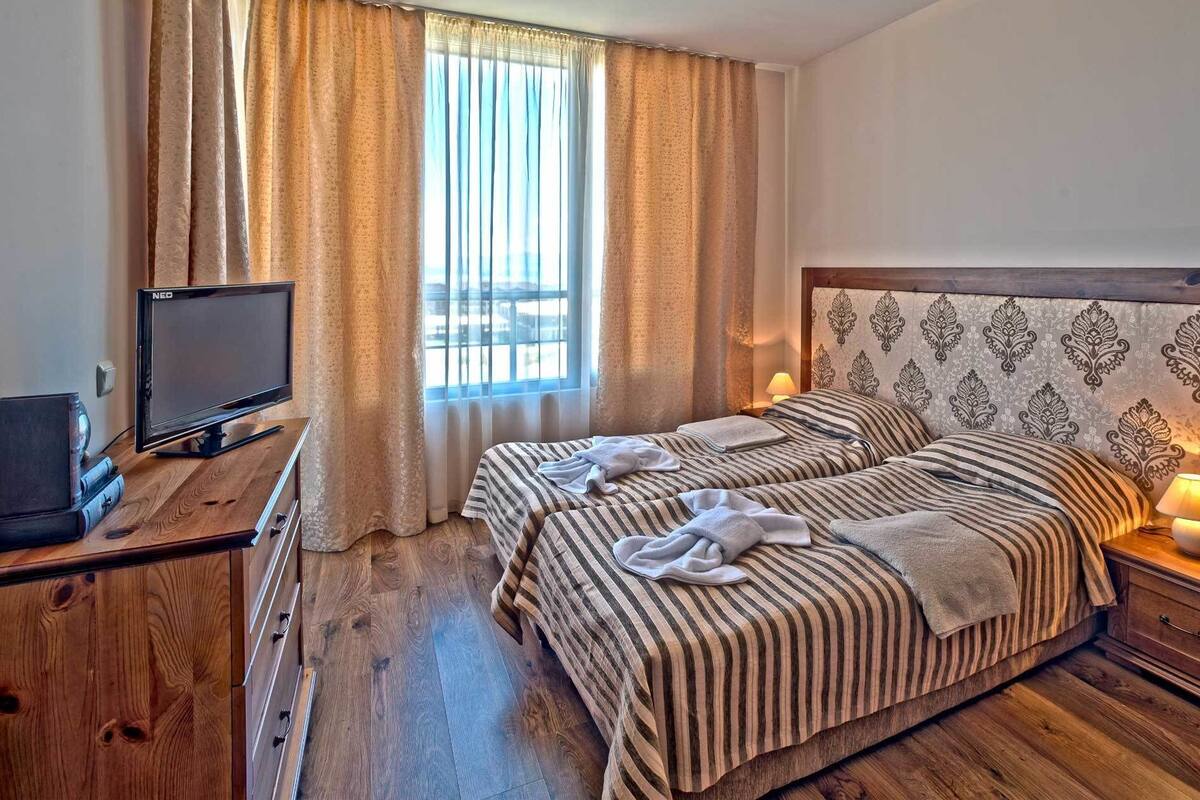 A hotel room with two beds, a TV, a desk with a computer, and a window with curtains.