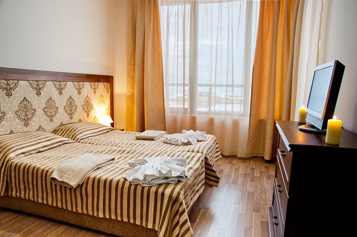Habitación de hotel con dos camas, un cabecero de madera, una mesita de noche con espejo y velas, y una ventana con cortinas.