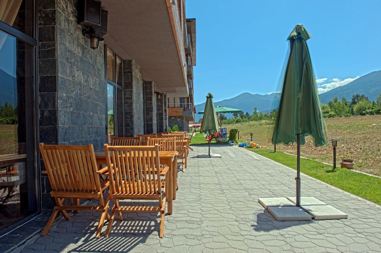 An outdoor seating area with wooden chairs and tables, a building with stone walls, and a view of mountains in the background.