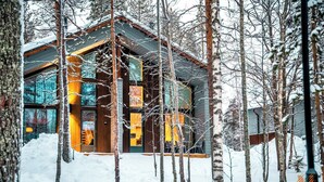 Une cabane en rondins moderne, dotée d’une grande porte et de grandes fenêtres en verre, entourée d’arbres couverts de neige.